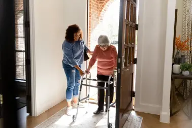 Eine Frau hilft einer älteren Frau durch die Haustür. Die ältere Frau nutzt eine Gehhilfe.